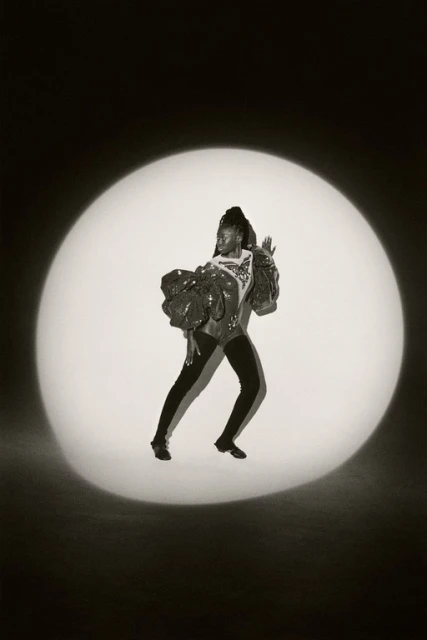 Woman in sequined top and black leggings strikes a pose in a spotlight against a dark backdrop, showcasing fashion and dance.