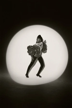 Woman in sequined top and black leggings strikes a pose in a spotlight against a dark backdrop, showcasing fashion and dance.