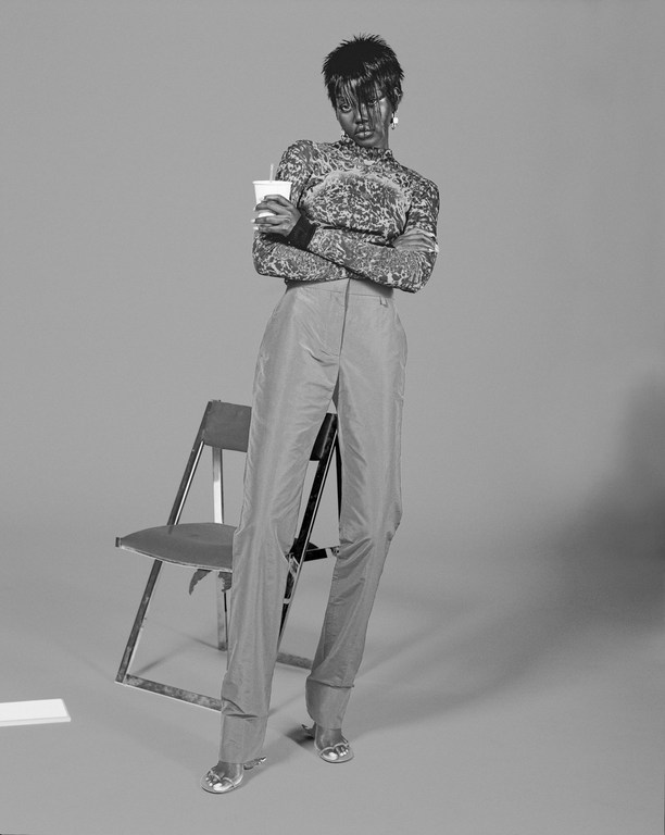 Black woman with short dark hair holding a cup, wearing patterned top and trousers, standing near a folding chair in a studio.
