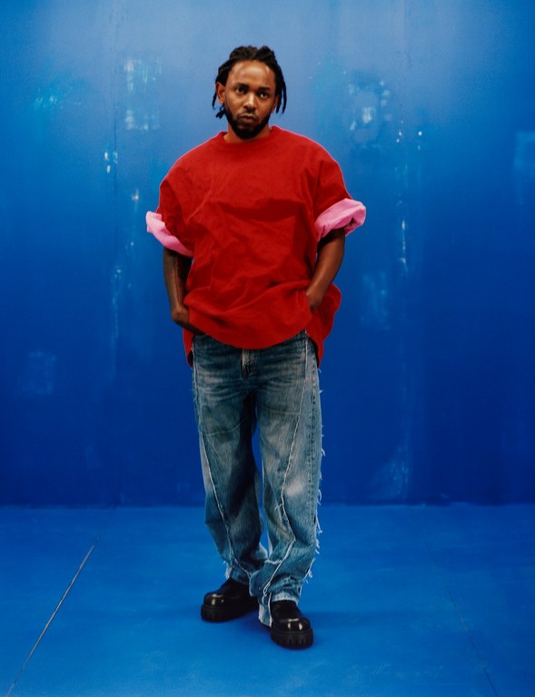 Portrait of Kendrick Lamar in a red t-shirt and distressed jeans against a blue backdrop, looking at the camera with a neutral expression.