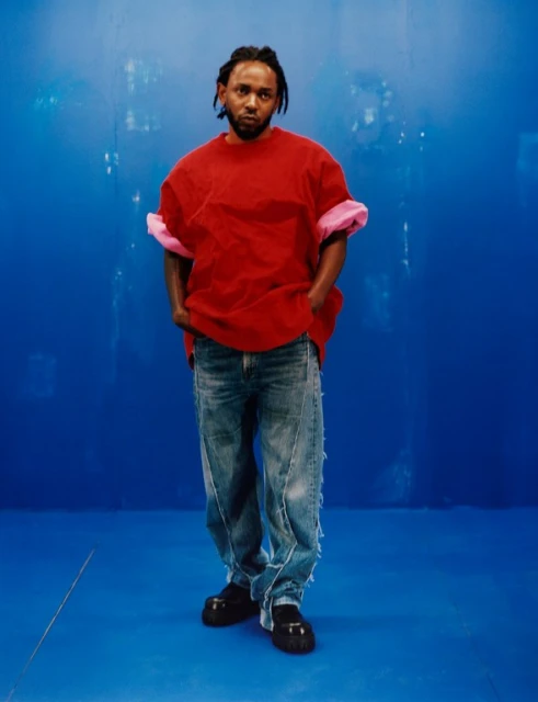 Portrait of Kendrick Lamar in a red t-shirt and distressed jeans against a blue backdrop, looking at the camera with a neutral expression.