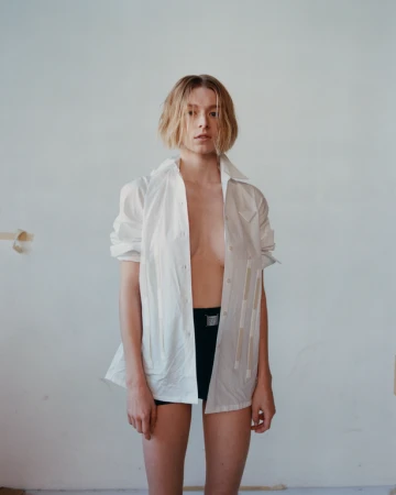 Woman with short blonde hair wearing an unbuttoned white shirt and dark shorts against a plain background