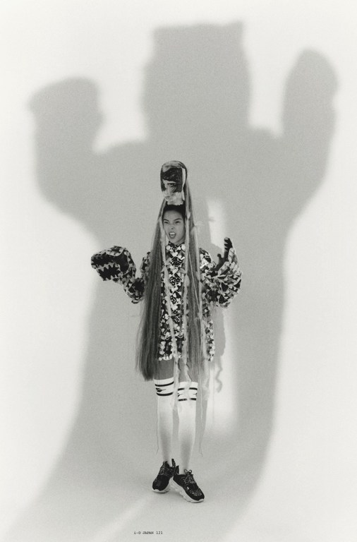 A woman poses in an avant-garde outfit with a patterned jacket, white socks, and black sneakers, standing in front of a plain background.