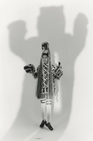 A woman poses in an avant-garde outfit with a patterned jacket, white socks, and black sneakers, standing in front of a plain background.
