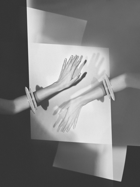 Monochrome art shot of a pair of arms emerging through white paper with silver bracelets, with hands extended upward in a studio.