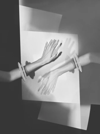 Monochrome art shot of a pair of arms emerging through white paper with silver bracelets, with hands extended upward in a studio.