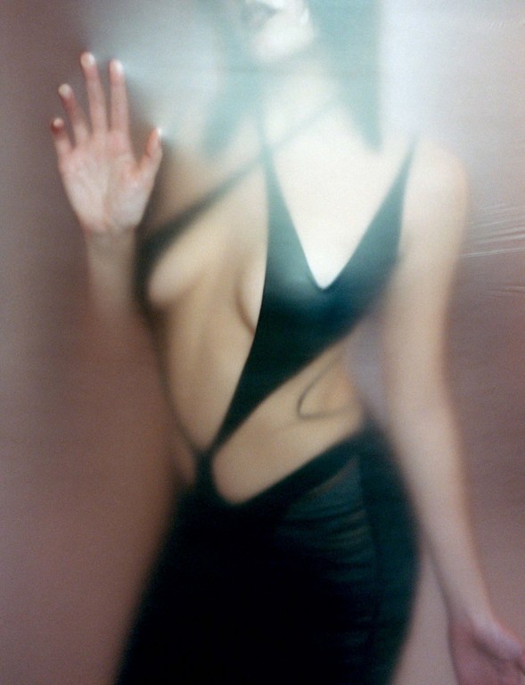 Woman in a black dress poses behind a textured semi-transparent glass, obscuring the image with an ethereal, diffused light effect.