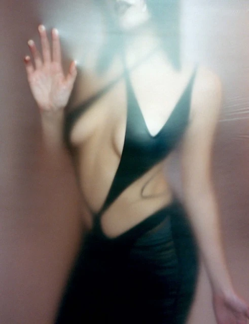 Woman in a black dress poses behind a textured semi-transparent glass, obscuring the image with an ethereal, diffused light effect.