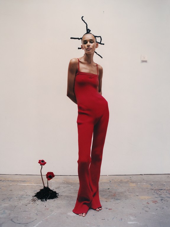 Fashion shot of a slender model in a red jumpsuit, posing with a blank wall backdrop and red flowers at her feet for a studio look.