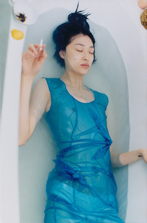 Woman in bathtub wearing blue dress and holding a cigarette, surrounded by water with floating lemon, eggs, and other objects.