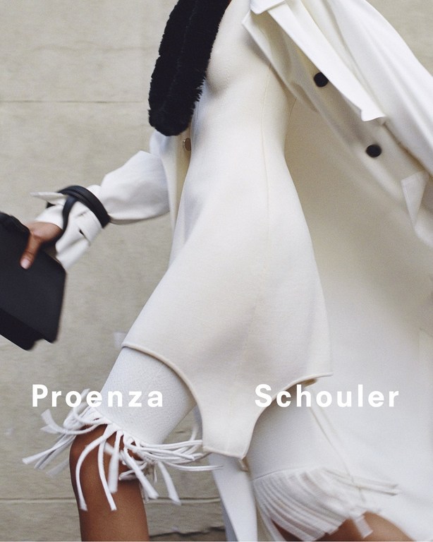 Fashion editorial shot of a person in a white dress and coat, holding a black case, walking against a neutral urban background.
