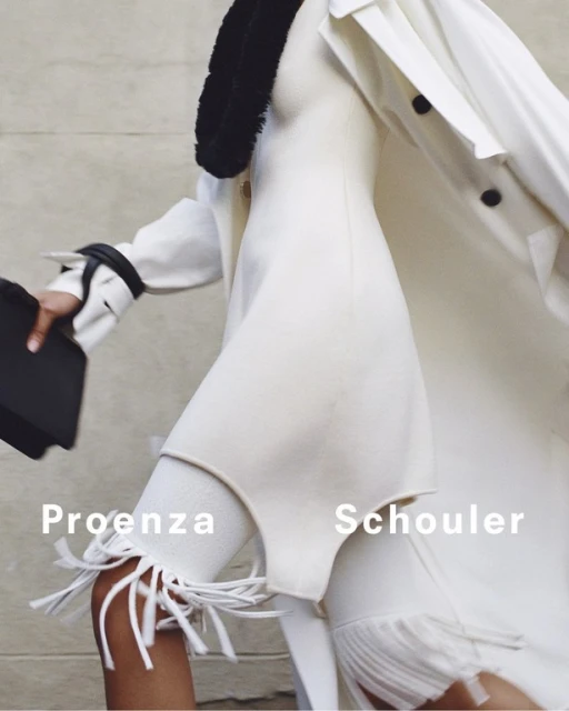 Fashion editorial shot of a person in a white dress and coat, holding a black case, walking against a neutral urban background.