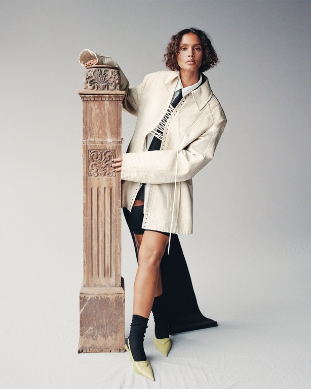 Woman poses in a white coat and black dress with a decorative pillar indoors, showcasing high-fashion style and neutral backdrop.