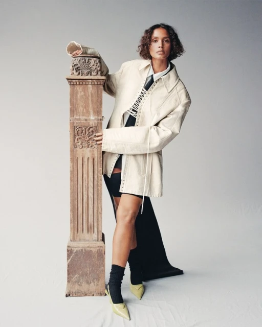 Woman poses in a white coat and black dress with a decorative pillar indoors, showcasing high-fashion style and neutral backdrop.