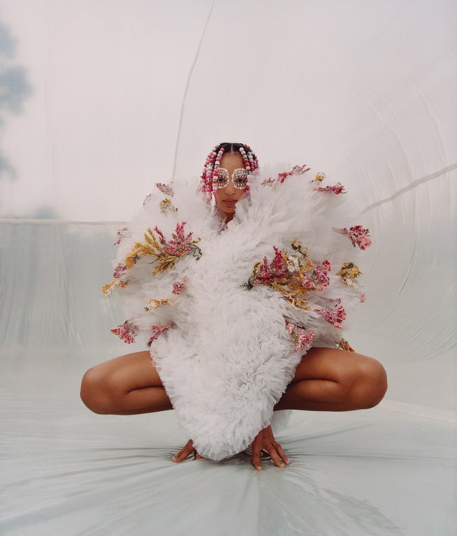Person in a white floral dress with pink-braided hair and vintage sunglasses poses in a bright studio, crouching, against a light background.