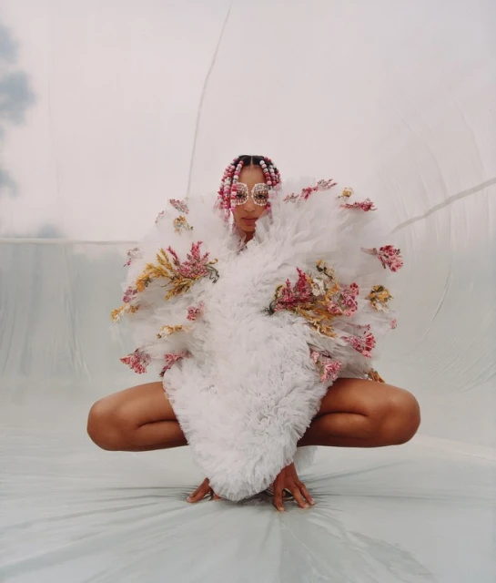 Person in a white floral dress with pink-braided hair and vintage sunglasses poses in a bright studio, crouching, against a light background.