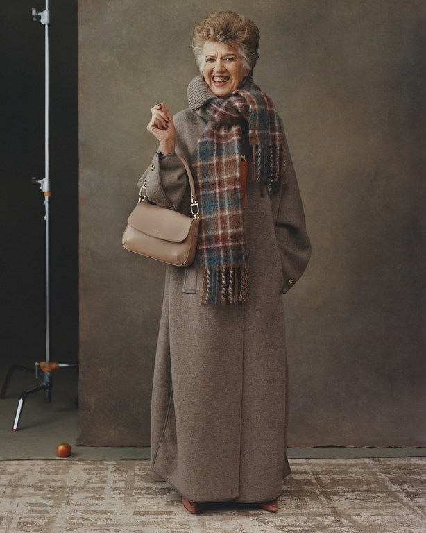 An elderly woman smiles while posing in a long brown coat, brown purse, and a multi-colored plaid scarf in a photo studio.
