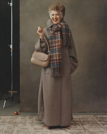 An elderly woman smiles while posing in a long brown coat, brown purse, and a multi-colored plaid scarf in a photo studio.