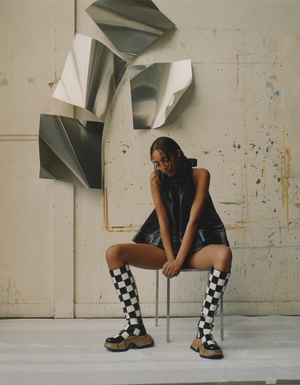 A woman sits on a chair in a photo studio wearing a black sleeveless dress and checkerboard print knee-high boots in a portrait.