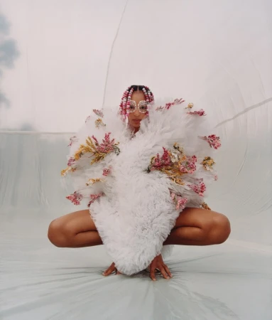 A person crouching down wearing a voluminous white textured garment adorned with flowers and beaded eyewear.