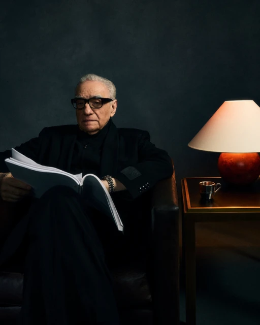 Portrait of an elderly man with glasses, wearing a dark coat, sitting in a leather chair while reading a book in a dimly lit room.