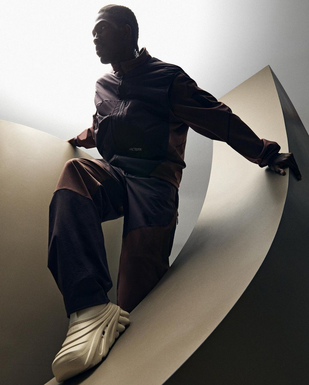 Dark-skinned male model in brown athletic wear poses on a curved structure in a low-angle shot with light flooding the background.