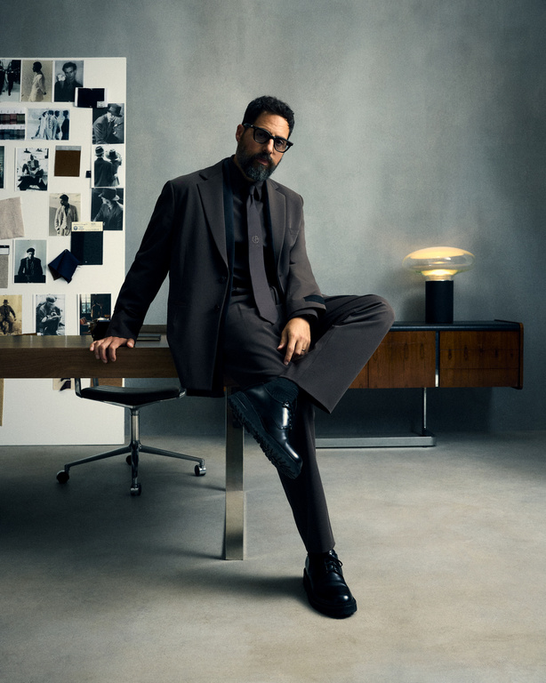 Man in a brown suit sits on a desk in a neutral-toned room with a moodboard and lamp, exuding a poised and professional demeanor.