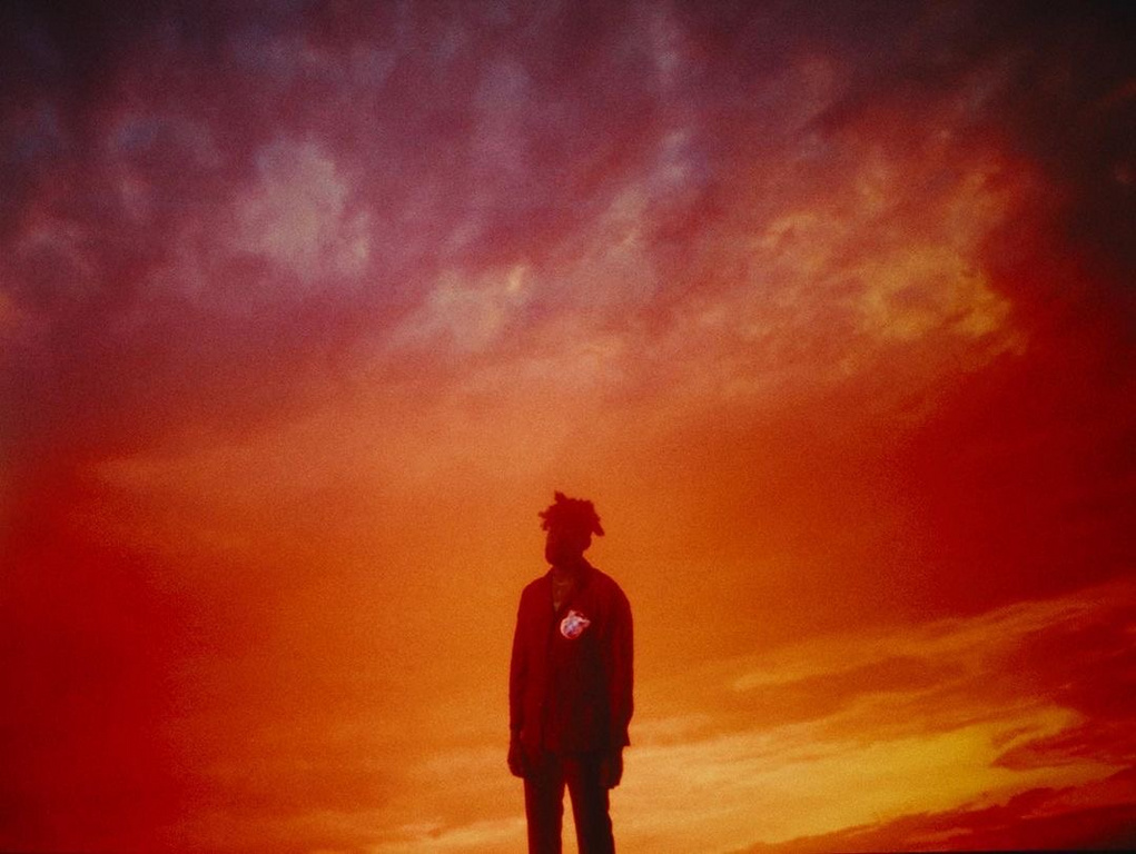 Silhouette of a person standing against a fiery orange and red sunset sky filled with dramatic clouds, creating a warm and intense atmosphere.