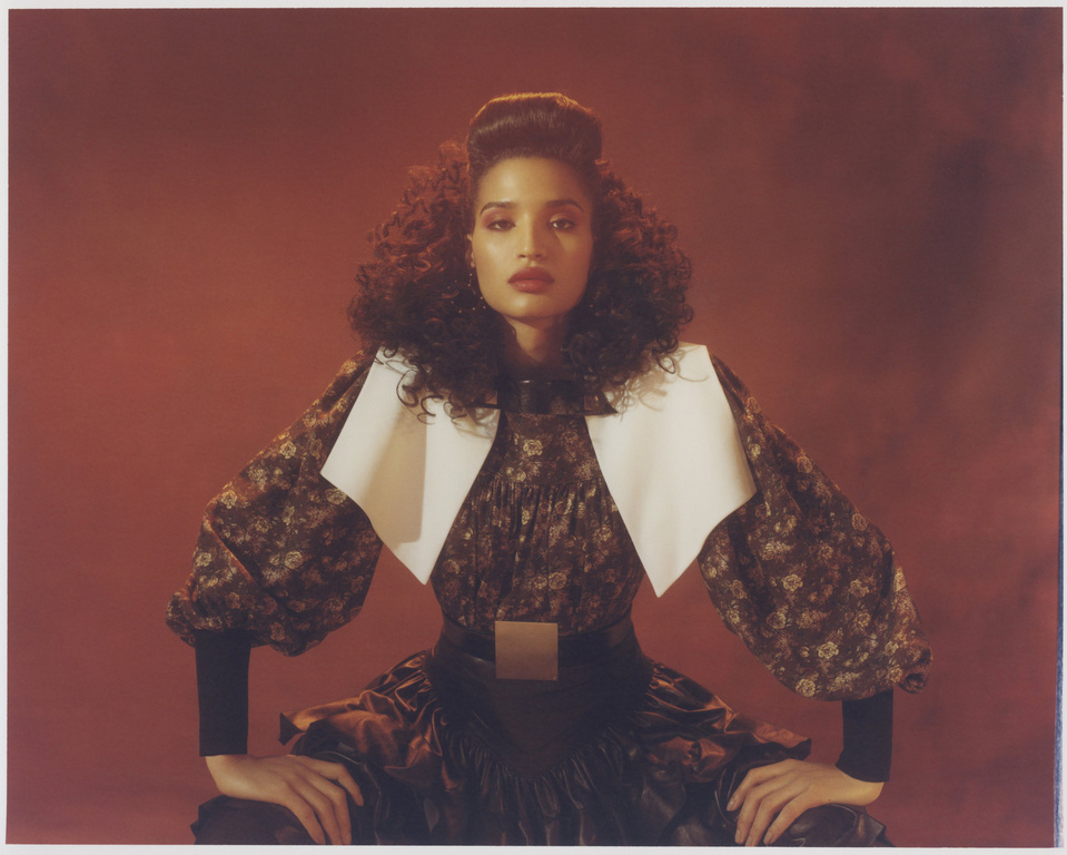 A woman poses indoors in a vintage-inspired floral dress and layered collar, captured in a portrait with moody lighting and a red background.