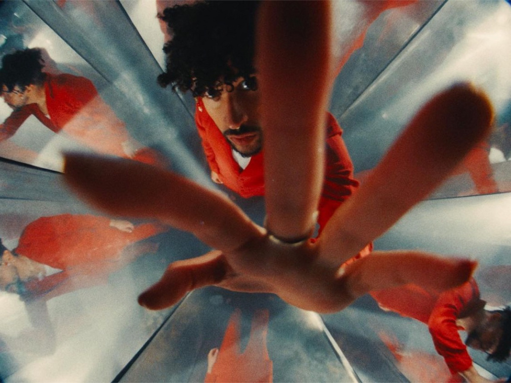 Low-angle shot of a man in red jumpsuit with curly hair reflected in mirrors creating a kaleidoscopic effect and distorted perspective.