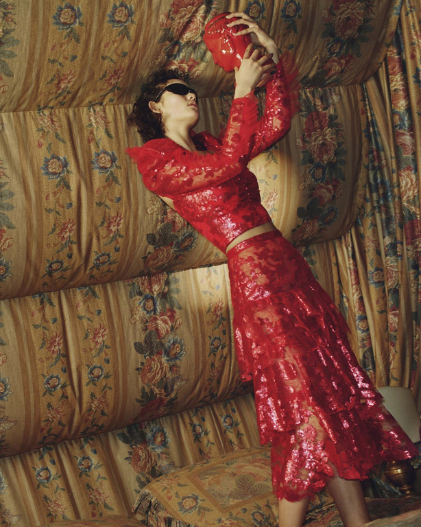 A person in red lace clothing poses indoors with a red, abstract, vase-like object, against a floral-patterned cushioned wall.