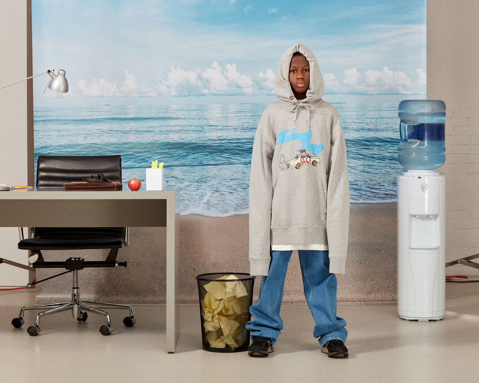 Young boy in a grey hoodie and blue jeans stands in an office space with a beach scene backdrop, featuring an office desk and water cooler.