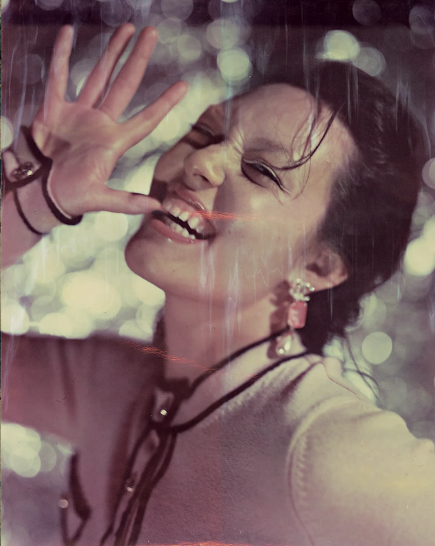 Woman with brown hair smiles in an eye-level, indoor shot, wearing jewelry, a light-colored top with a dark collar, and light raindrops.