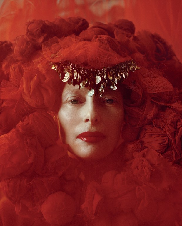 Portrait of a woman with fair skin and red lipstick, adorned with a red veil and an ornate metal headpiece with coin-like details.