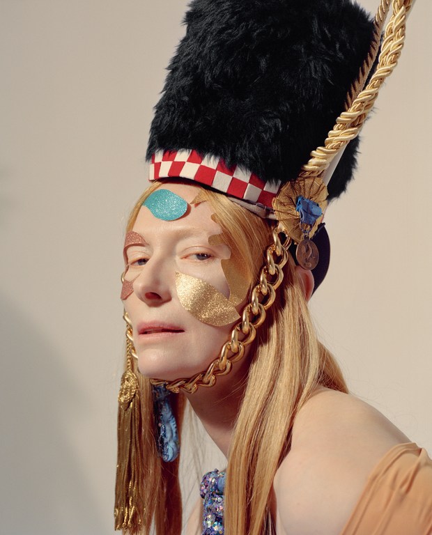 Portrait of a fair-skinned adult with red hair wearing an elaborate fur hat and decorative face jewelry in a photo studio.