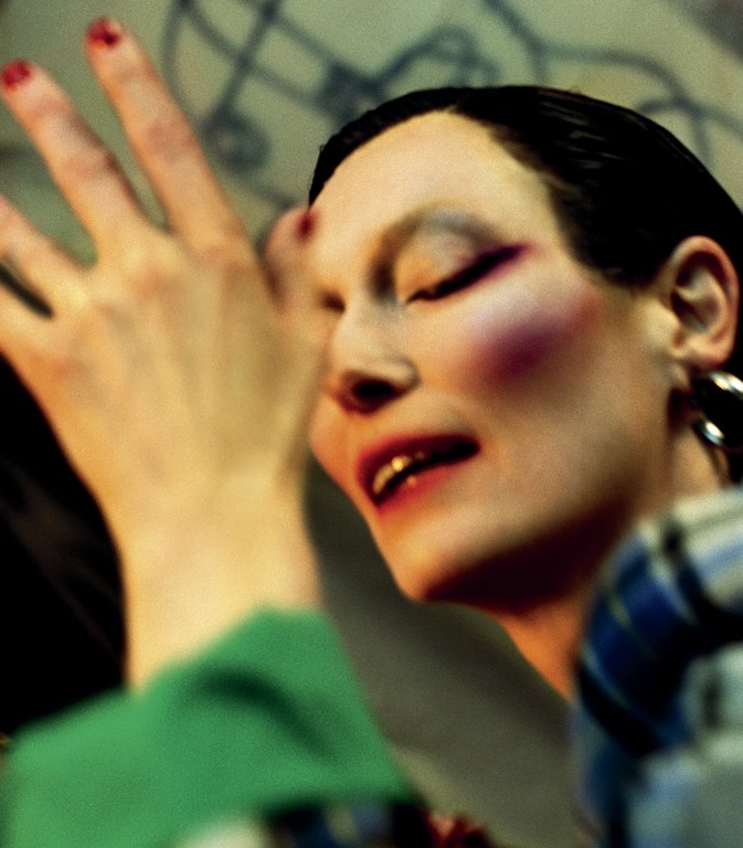 Adult female face with bold makeup and large hoop earrings in soft lighting with dark hair and hand partially visible in the frame.
