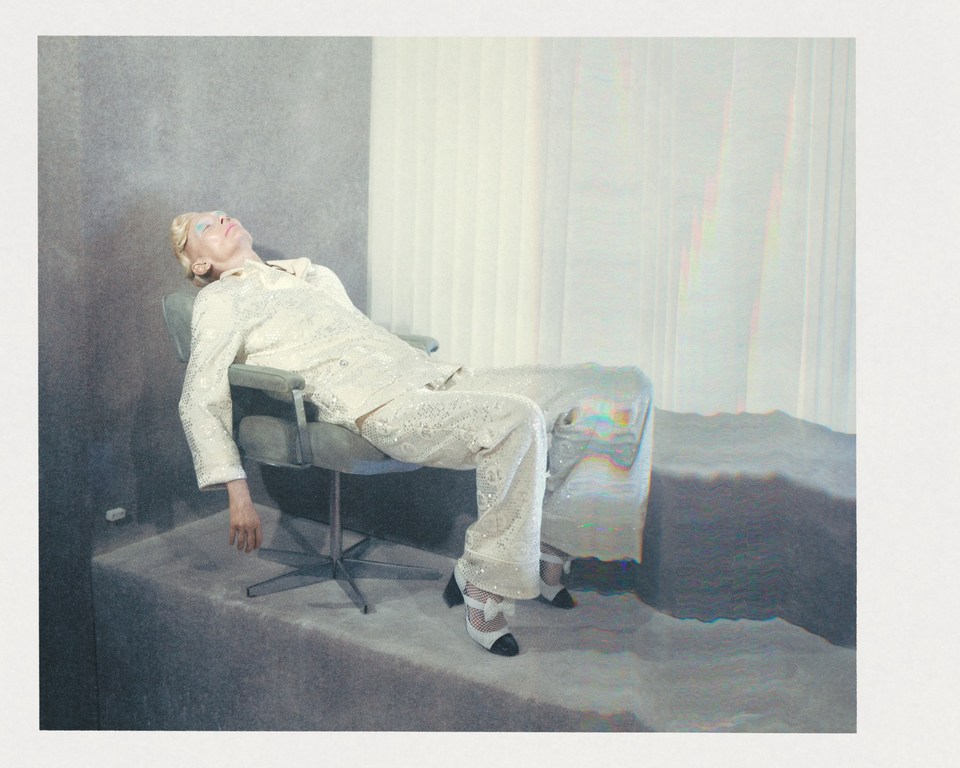 Elegant adult woman in a patterned suit reclines in a modern chair in a minimalist setting with a concrete platform and sheer curtains.