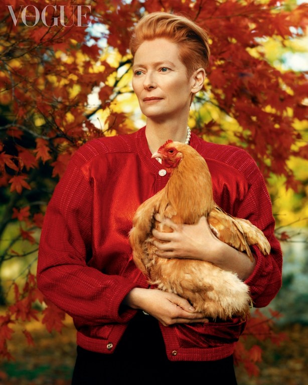 Elegant woman in red jacket holds a light brown chicken outdoors in a maple-leaf setting, graced by the iconic 'Vogue' logo.