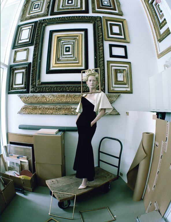 Elegant woman in a gown stands on a wooden platform, framed by picture frames and white walls. Art studio setting with natural light.
