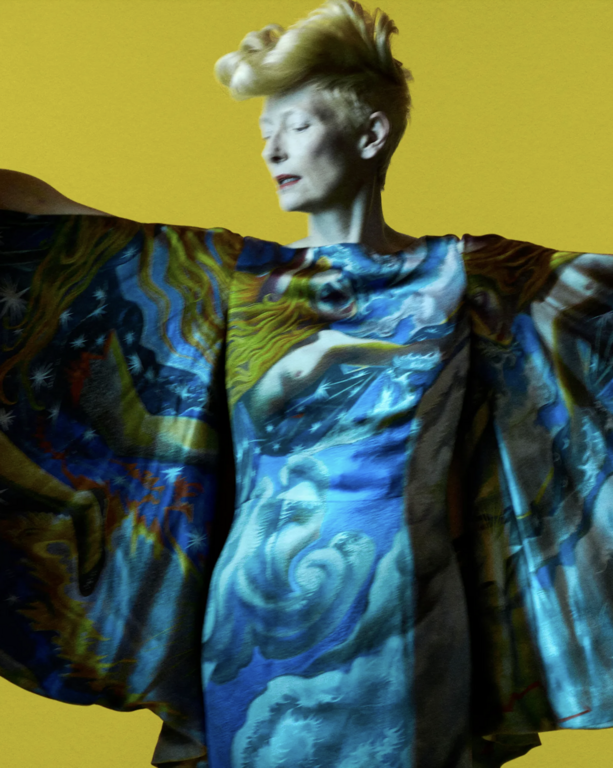 Adult female with blonde hair in a patterned, flowing dress posing in front of a yellow studio background with arms outstretched.