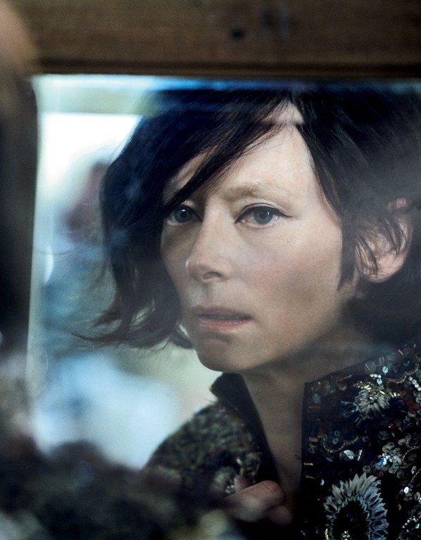 Close-up portrait of a woman with dark hair and light skin, wearing an embroidered jacket, looking towards a reflection in a mirror or glass.