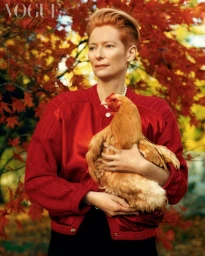 Tilda Swinton with red hair wearing a red jacket holding a chicken outdoors with autumn foliage