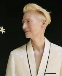 Tilda Swinton with blonde hair wearing a patterned cream jacket against a black background, styled by Jerry Stafford