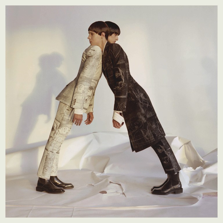 Two young men stand back-to-back in patterned suits, leaning against each other, on a crumpled white paper backdrop in a studio setting.