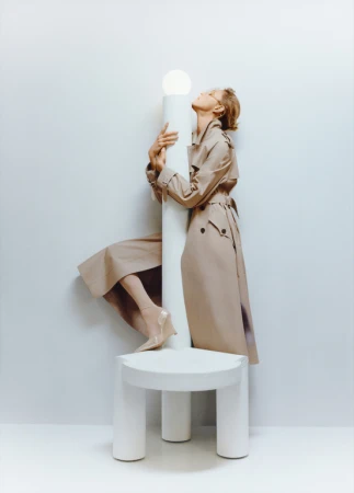 Woman in trench coat leans head back against white column on round white table in white studio. Light skin and blonde hair.