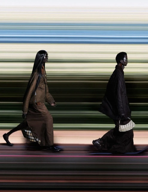 Two adult fashion models walk side-by-side in a studio wearing brown full-length outfits against a blurred background with horizontal bands.