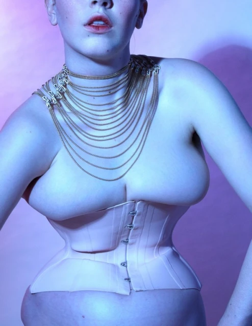 Elegant portrait of a woman wearing a pale corset and gold chain necklace against a soft pink background in a photo studio setting.