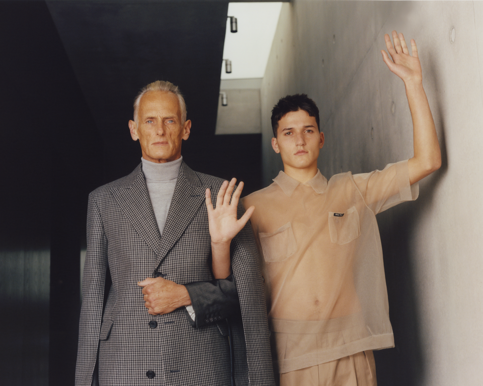 Older and younger adult males in an indoor setting. The men are wearing fashionable clothing, gesturing with their hands, and facing forward.