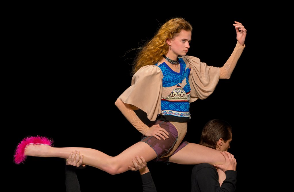 Fashion model on a catwalk, performing splits while being supported by two male figures against a black backdrop during a runway show.