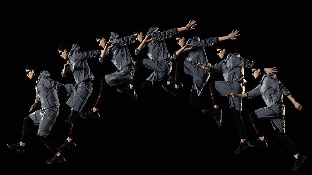 Man in a grey running suit performing a sequence of jumping jacks against a black background, captured with a long exposure technique.